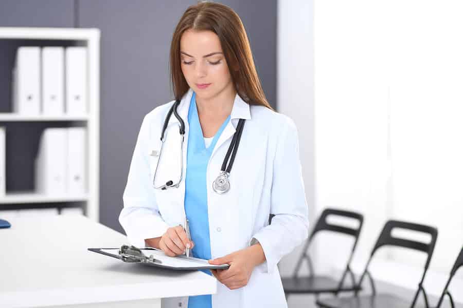 Doctor woman standing at work. Portrait of female physician filling up medical form while standing near reception desk at clinic or emergency hospital. Medicine and healthcare concept